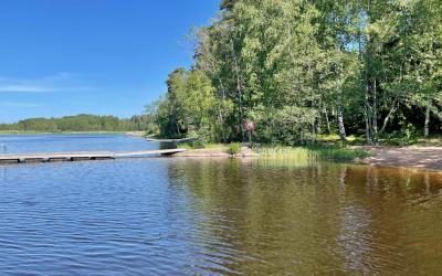 kesäisessä maisemassa puulaituri ja hiekkaranta, tausalla lehtipuita