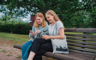Kaksi teini-ikäistä tyttöä istuvat vierekkäin ruskealla puiston penkillä ja molemmat katsovat hymyillen puhelimiaan. Taustalla näkyy kesäisen vihreää puistomaisemaa.