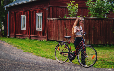 Nuori nainen kävelee ja taluttaa samalla pyörää. Taustalla punainen vanha rakennus.