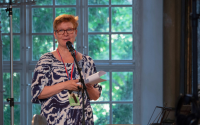 Nainen pitää puhetta, taustalla vanhan kirkon iso ikkuna ja vehreän kesäinen maisema.