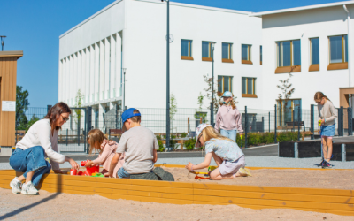 Aikuinen ja lapsia leikkimässä hiukkalaatikolla aurinkoisena kesäpäivänä.