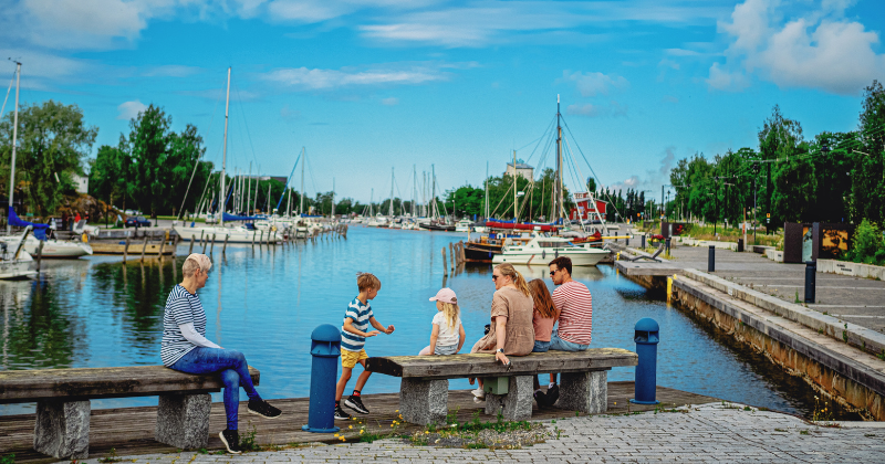 2 aikuista ja 3 lasta istuvat Uudenkaupungin kaupunginlahden rannalla penkillä. Taustalla sininen taivas, merta ja veneitä.