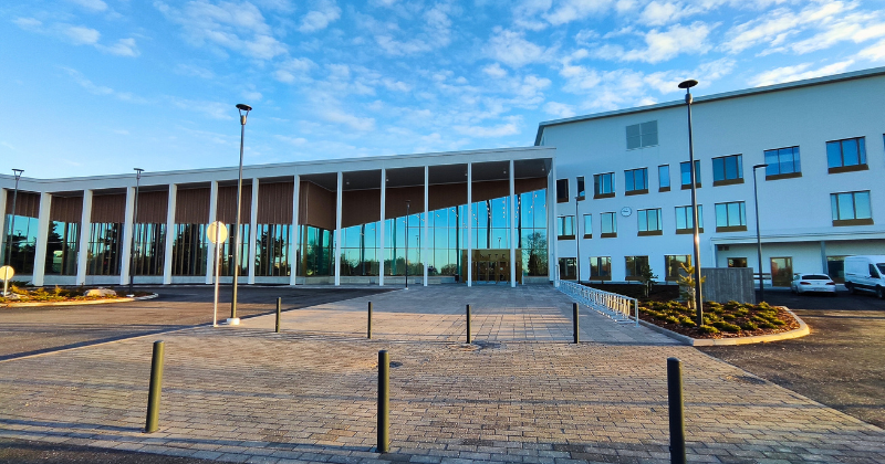 Edessä uusi moderni rakennus, jossa valkoista rapattua pintaa ja toisessa sivussa isot lasiseinät vinoine kattoineen.