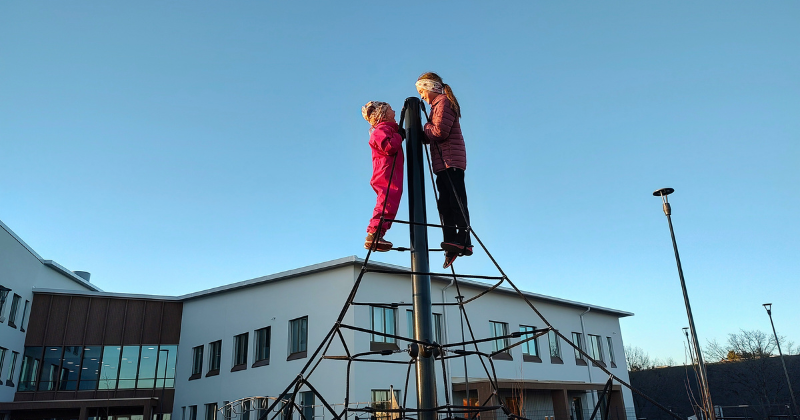 Kaksi lasta seisoo rakennuksen edessä olevassa lasten kiipeilytelineen huipulla. Taustalla vaaleahko koulurakennus ja sininen taivas.