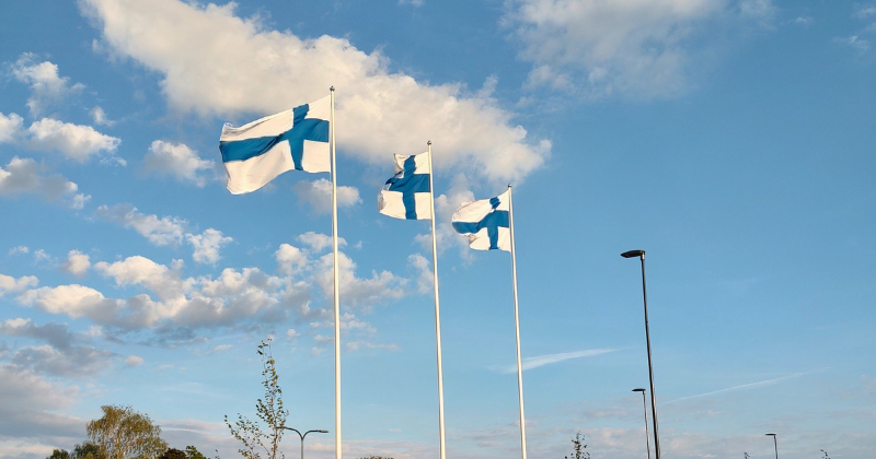 Kolme Suomen lippua liehuu aurinkoisessa ilmassa, sininen taivas pilvineen taustalla.