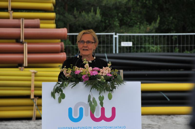 Nainen pitää puhetta puhujankorokkeelta. Korokkeessa kukkia ja taustalla muovisia rakennusputkia.