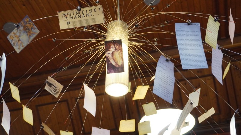 Lähikuva kirjaston valaisimesta. Valaisimessa on vaijereita, joihin on kiinnitetty papereita. Papereissa on kuvia ja tekstiä.